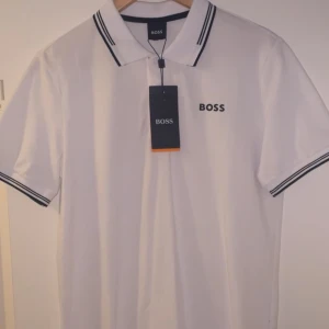 Vit pikétröja från HUGO BOSS - Säljer en stilren vit pikétröja från HUGO BOSS med klassisk krage och svarta detaljer. Tröjan har korta ärmar och en liten BOSS-logga på bröstet. Perfekt för en avslappnad men ändå snygg look.