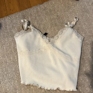 Vit ribbad linnetopp från H&M Divided - Säljer en söt vit ribbad linnetopp från H&M Divided. Toppen har spetsdetaljer vid halsringningen och axelbanden är knytbara. Perfekt för varma dagar eller som ett lager under en kofta.