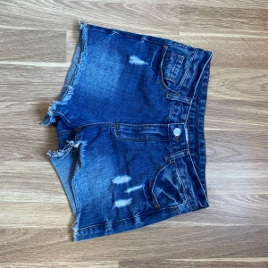 Blå jeansshorts från Lindex - Snygga blå jeansshorts från Lindex med fransiga kanter och slitna detaljer. Perfekta för en avslappnad stil. Shortsen har klassisk femficksdesign och knappgylf.