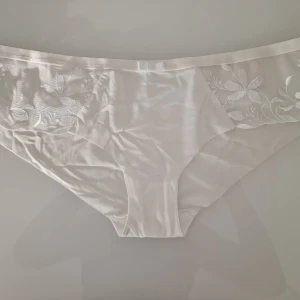 Vit spetsdetaljerad trosa från Triumph  - Romantisk vit trosa med vackra spetsdetaljer på sidorna. Trosan har ett blommigt mönster i spetsen som ger en elgant känsla. Endast tvättad och har sedan blivit liggandes i lådan.