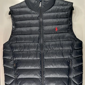 Svart väst från Polo Ralph Lauren - Snygg svart väst från Polo Ralph Lauren med röd broderad logga på bröstet. Perfekt för lager-på-lager under kyliga dagar och nu inför våren. Västen har en quiltad design som ger en stilren look. Den är helt ny, aldrig använd. Tags sitter på. Kontakta vid intresse. 