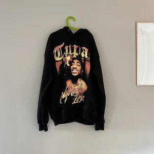 Svart Tupac hoodie från Pull&Bear - Säljer en svart hoodie från Pull&Bear med Tupac-tryck och texten 'California Love' på framsidan. Hoodien har en klassisk passform med långa ärmar och en bekväm huva. Perfekt för dig som älskar ikoniska musikmotiv!