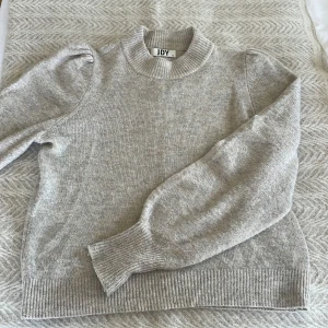 Grå stickad tröja från JDY - Säljer en stilren grå stickad tröja från JDY. Tröjan har långa ärmar med puffiga axlar och ribbade muddar vid ärmslut och nederkant. Perfekt för kyliga dagar!