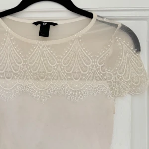Vit blus med spetsdetaljer från H&M - En elegant vit blus från H&M med vackra spetsdetaljer på axlar och överdel. Blusen har korta ärmar och en rund halsringning. 
