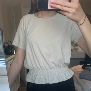 Beige topp med resårdetalj - Säljer en stilren beige topp med korta ärmar och resår i midjan som ger en fin volangeffekt. Perfekt för en avslappnad look. Passar bra till både jeans och kjol.