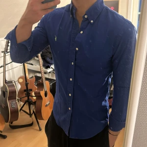 Blå skjorta från Ralph Lauren - Snygg blå skjorta från Ralph Lauren med klassisk krage och knappar framtill. Skjortan har en broderad logga i grönt på bröstet och långa ärmar. Perfekt för en stilren look.