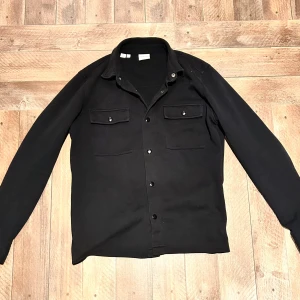 Svart overshirt från Selected Homme - Snygg svart overshirt från Selected Homme med långa ärmar och knappar framtill. Två praktiska bröstfickor med knäppning ger en stilren look. Perfekt för lager-på-lager-stil.