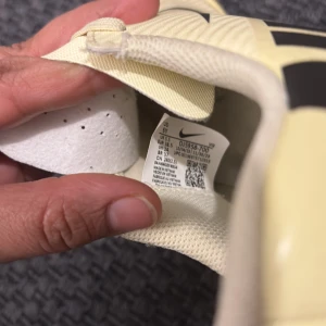 Beige fotbollsskor från Nike - Snygga beige fotbollsskor från Nike med svarta detaljer och snörning. Skorna har en klassisk design med dobbar för bra grepp på planen. Perfekta för fotbollsträning och matcher.
