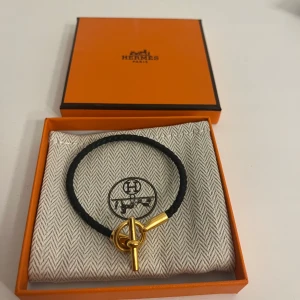 Svart armband från Hermès - Elegant svart armband från Hermès med en enkel design. Armbandet har en flätad rem och en stilren guldfärgad låsdetalj med Hermès-logotypen ingraverad. Perfekt för att ge en lyxig touch till din stil.
