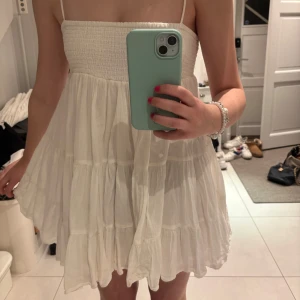 Vit ärmlös klänning - Säljer en söt vit klänning med smala axelband och smockad överdel. Kjolen är luftig och har flera lager som ger en fin volym. Perfekt för varma dagar!