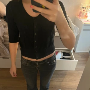 Svart kofta från Lindex - Säljer en stilren svart kofta från Lindex med knappar framtill. Koftan har trekvartsärmar och en ribbad kant vid ärmslut och nederkant. Perfekt för en enkel och snygg look.