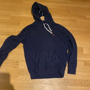 Mörkblå hoodie från Polo Ralph Lauren - Säljer en snygg mörkblå hoodie från Polo Ralph Lauren med en klassisk känguruficka och justerbar huva med vita snören. Den har en liten röd logotyp på bröstet. Använd några gånger, är i ett bra skick 