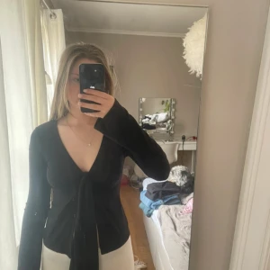 Svart omlottopp från Gina Tricot - Snygg svart omlottopp från Gina Tricot med knytning framtill. Toppen har långa ärmar och en djup v-ringning, perfekt för en stilren look. Passar bra till både jeans och kjol.