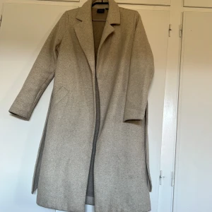 Beige kappa från Veromoda  - Elegant beige kappa från Veromoda med klassisk krage och långa ärmar. Perfekt för höst och vår, med en stilren design som passar till många tillfällen. Kappan har en rak passform och är tillverkad i ett mjukt material. 