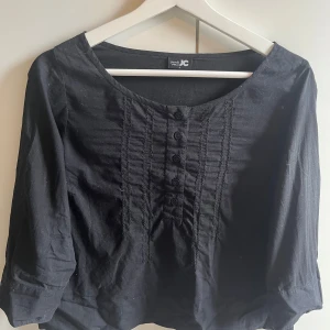 Svart blus från JC - Snygg svart blus från JC med trekvartsärm och knappar framtill. Perfekt för en stilren look. Blusen har en rund halsringning och dekorativa sömmar som ger en extra touch.