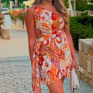 Färgglad mönstrad klänning - Snygg och färgglad klänning med ett livfullt mönster i rött, orange och vitt. Klänningen är ärmlös och har en knytning i midjan som ger en fin siluett. Perfekt för sommarens alla tillfällen!