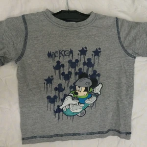 Grå Disney T-shirt med Musse Pigg 4 år - Säljer en grå T-shirt från Disney med Musse Pigg-tryck. T-shirten har korta ärmar och är gjord av en mjuk bomullsblandning. Perfekt för alla små Musse Pigg-fans! 🐭 88% bomull, 12% polyester