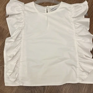 Vit blus med volangdetaljer från Almadirect - Säljer en stilren vit blus från Ginatricot med volangdetaljer längs sidorna. Blusen har korta ärmar och en rund halsringning. Perfekt för en elegant look. Kontakta mig för mer info!