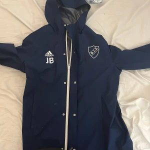 Blå regnjacka från Adidas - Säljer en blå regnjacka från Adidas med DIF-logga och initialerna JB på framsidan. Jackan har en huva och dragkedja framtill. På baksidan står det 'Stadium Up For Match'. Perfekt för regniga dagar! Pris kan diskuteras 