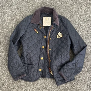 Mörkblå quiltad jacka från Morris - Säljer en snygg mörkblå quiltad jacka från Morris. Jackan har en klassisk design med bruna detaljer och guldknappar. Perfekt för höst och vår med sin stilrena look och bekväma passform.