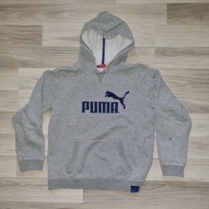 Grå hoodie från Puma - Säljer en grå hoodie från Puma med blå logga på bröstet. Tröjan har en klassisk känguruficka och ribbade muddar vid ärmslut (något nöta/frasiga) och nederkant. Perfekt för en avslappnad stil.