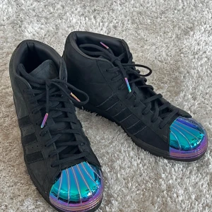 Svarta Adidas sneakers med metall tå - Oanvända snygga svarta Adidas sneakers med en unik metall tå i regnbågsfärger. Skorna har klassisk snörning och de ikoniska tre ränderna på sidan. Perfekta för dig som vill sticka ut med en cool och modern stil.
