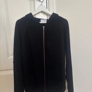 Svart hoodie i kashmir  - Svart hoodie från Davida i mjuk kashmir. Tröjan har dragkedja framtill, huva med snörning och två fickor. Perfekt för dig som gillar en clean och stilren look med lyxig känsla. Passformen är normal och ärmarna är långa. Aldrig använt så tagsen är fortfarande på skriv om ni har några funderingar!