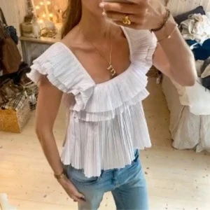 Vit volangblus från Zara - Supersöt vit blus från Zara med breda volangärmar och plisserade detaljer. Toppen är ärmlös och har flera lager volanger som ger en luftig och romantisk känsla. Perfekt att styla med jeans eller kjol för en trendig look.