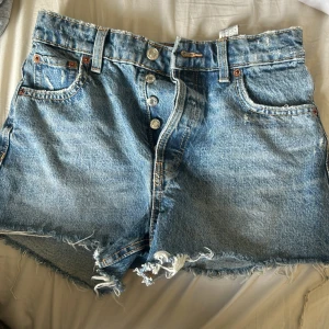 jeansshorts från Zara - Snygga blå jeansshorts från Zara med medelmidja, råa och fransiga benslut samt klassisk femficksdesign. Shortsen har knappar framtill och är tillverkade i slitstarkt denim. Perfekta för en chill sommarlook.
