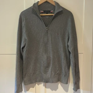 Grå stickad kofta Tommy Hilfiger - Snygg grå stickad kofta från Tommy Hilfiger med dragkedja framtill och liten broderad logga på bröstet. Modellen har hög krage och långa ärmar, perfekt för lager-på-lager. Klassisk och stilren design som funkar året runt.