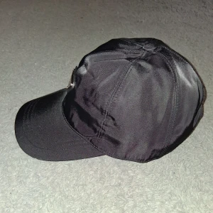 Svart keps från Prada i nylon - Snygg svart keps från Prada i återvunnen nylon med klassisk böjd skärm och ikonisk metallogo framtill. Kepsen har justerbar rem baktill och stilren design som passar perfekt till streetwear eller en clean look.