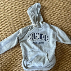 Grå hoodie med California-tryck - Snygg grå hoodie med stor huva och snörning. Framtill står det 'CALIFORNIA SAN FRANCISCO' i mörkblå text. Klassisk känguruficka och mjukt material, perfekt för chill dagar eller när du vill ha en avslappnad look.
