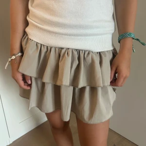 Beige volangkjol från Neo Noir - Supersöt beige kortkjol från Neo Noir med dubbla volanger och elastisk midja. Kjolen har en flowy passform och är perfekt för dig som gillar en feminin och trendig look. Lätt att matcha med både stickade tröjor och linnen.