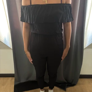 Svart off shoulder byxdress - Snygg svart byxdress med off shoulder-överdel och smala ben. Mjuk och lätt i materialet, perfekt för dig som gillar en stilren look. Resår i midjan för skön passform och trendig volang. 