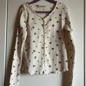 Vit ribbad tröja med blommigt mönster - Supersöt vit ribbad tröja med svarta små blommor och knappar framtill. Långärmad och med lätt vågig kant nertill.