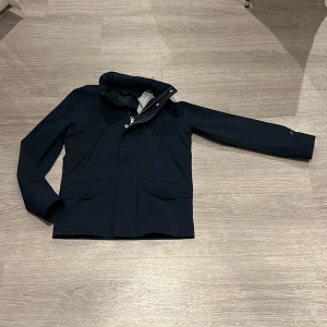 Mörkblå jacka från J.Lindeberg - Snygg mörkblå jacka från J.Lindeberg i minimalistisk design. Jackan har vattentät dragkedja, två stora fickor framtill och hög krage. Perfekt för blöta dagar och stilren look. Tillverkad i syntetmaterial och har långa ärmar. Luva tillgår och ligger i kragen . Helt oanvänd och i nytt skick!