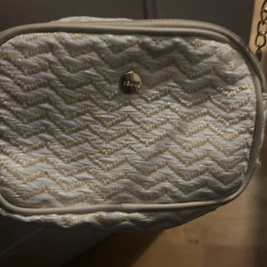 Vit axelväska med kedja från Lindex - Säljer en söt vit axelväska från Lindex med beige zigzag-mönster och guldfärgad kedjerem. Väskan har en rundad form och är tillverkad i ett flätat material som ger en fräsch och somrig vibe. Perfekt för att lyfta din outfit med en trendig detalj.