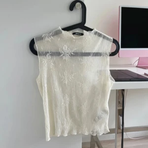 Viral mesh topp från Gina Tricot - Säljer en vit ärmlös mesh topp från Gina Tricot med blommigt mönster och lätt vågig struktur. Toppen är transparent och har en hög hals.