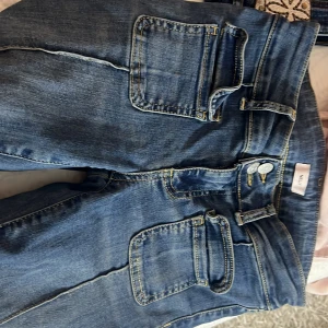 Blå jeans med stora fickor från Nelly - Snygga blå jeans från Nelly med markerade sömmar och stora fyrkantiga fickor framtill. Jeansen har dubbla silverknappar i midjan och klassisk denimlook. Perfekta för dig som gillar en cool och avslappnad stil.