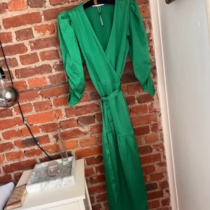 Grön omlottklänning med puffärm - Oanvänd snygg lång grön omlottklänning med knyt i midjan och puffiga långärmar. Klänningen har v-ringning och ett lätt glansigt tyg som ger en lyxig känsla. Perfekt för dig som vill sticka ut med färg och stil. Passar även medium utan problem. Nypris 800kr. Prislappen är ej kvar men den är ej använd.