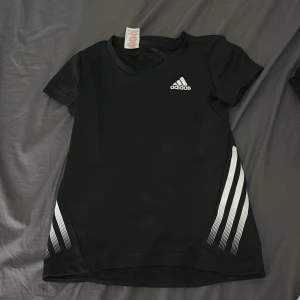 Svart Adidas t-shirt med vita ränder - Svart t-shirt från Adidas med klassiska vita ränder på sidorna och Adidas-logga på bröstet. Gjord i mjuk polyester, perfekt för träning eller sport. Kortärmad och med rund hals. Snygg och sportig stil som alltid funkar. 