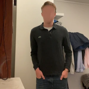 Mörkgrå pikétröja från Lacoste XS - Snygg mörkgrå pikétröja från Lacoste i klassisk modell med krage och knappar. Ikoniska gröna krokodilloggan broderad på bröstet. Tillverkad i 100% bomull för en skön och avslappnad känsla. Perfekt till jeans eller chinos.