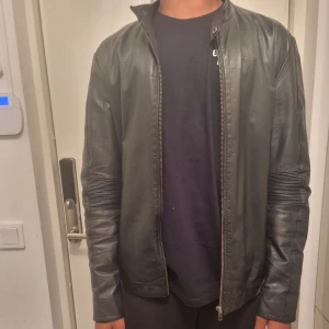 Svart skinnjacka med dragkedja -  Äkta Snygg svart skinnjacka med coola ribbade detaljer på ärmar och rygg. Jackan har en rak passform, dragkedja framtill och ståkrage. Perfekt för dig som vill ha en stilren och edgy look. Passar till det mesta och ger en grym vibe.         Är 185 på bild