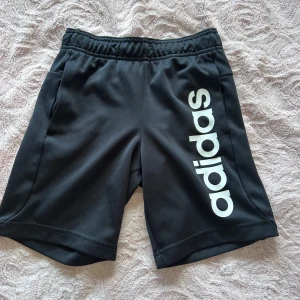Svarta Adidas shorts med logga - Svarta sportiga shorts från Adidas med stor vit logga längs ena benet. De har elastisk midja och är gjorda i ett mjukt, lätt polyestermaterial som andas. Perfekta för träning eller chill dagar. Klassisk och clean look som funkar till det mesta. De har en ficka på varje sida fram.
