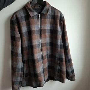 Rutig overshirt från Clean Cut Copenhagen - Snygg overshirt i brunt, grått och svart från Clean Cut Copenhagen. Jackan har ett rutigt mönster, dragkedja framtill och klassisk krage. Perfekt lager-på-lager-plagg för höst och vår. Tillverkad i mjukt material som känns skönt mot huden. Ny pris: 990kr. 