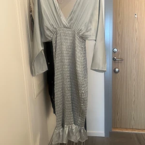 Ljusgrå omlottklänning med volang - Supersnygg ljusgrå långklänning med omlottliv, v-ringning och långa ärmar. Kjolen är smockad och avslutas med en volangkant. Funkar för S och M eftersom den är stretchig.