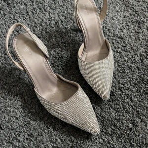 Glittriga pumps med spetsig tå - Snygga pumps från Gina Tricot med spetsig tå och öppen häl. Skorna är täckta av glittrande stenar och har en elegant, smal klack. Färgen är silver och materialet är syntet. Några stenar har ramlat av men inget som syns på håll utan bara på nära håll!