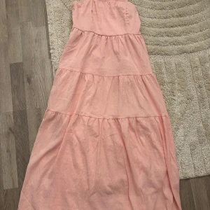 Ljusrosa axelbandslös långklänning - Säljer en söt ljusrosa långklänning med volangiga lager och axelbandslös design. Perfekt för varma dagar och somriga vibes. Klänningen har en luftig passform och är tillverkad i ett lätt material som känns skönt mot huden.