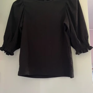 Svart blus med puffärm från Lindex - Svart blus från Lindex i storlek XS med puffiga trekvartsärmar och smockdetaljer vid ärmslut. Toppens tyg har en lätt struktur och rund halsringning. Perfekt för dig som gillar stilrena och trendiga plagg med lite volym i ärmarna. 🩵🩵🩵