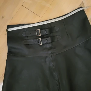 Skinnkjol med spännen, mörkblå  - Snygg kort skinnkjol med två coola spännen i midjan. Kjolen har en lätt A-linjeform och är perfekt för dig som gillar en edgy stil. Materialet är slätt och ger en lyxig känsla. Passar perfekt till boots eller en oversized tröja.